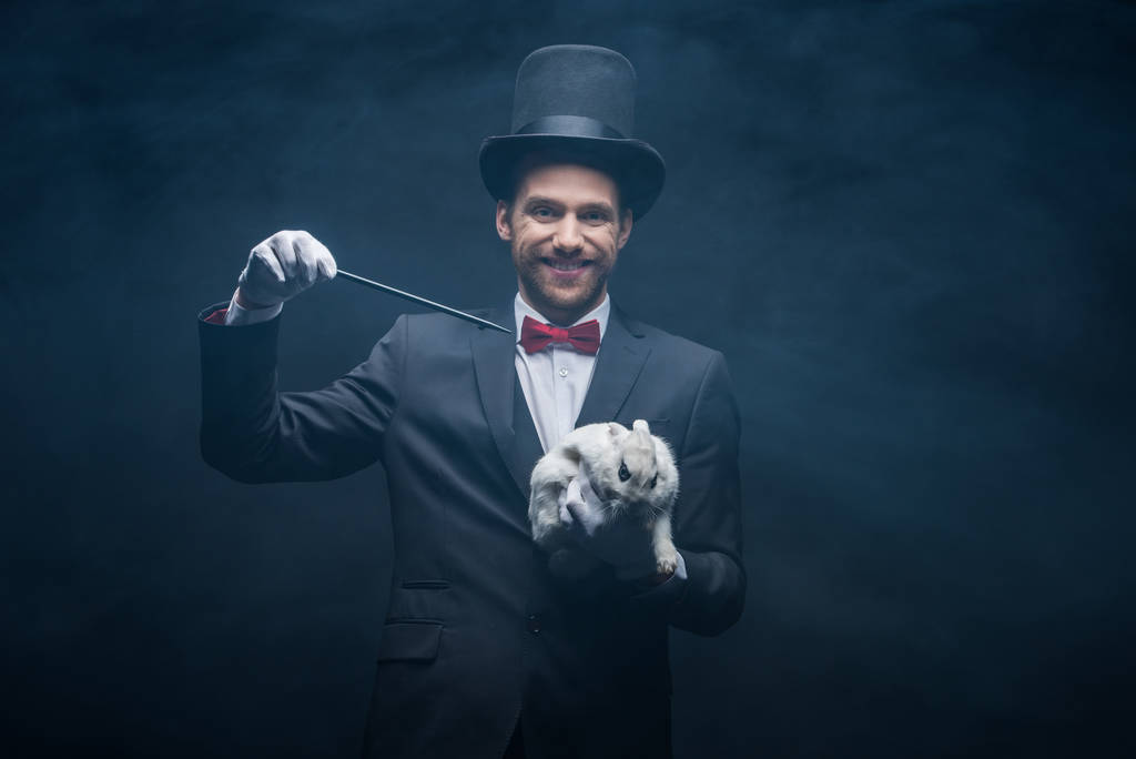 cheerful magician in suit showing trick with wand and white rabbit in hat, dark room with smoke - Photo, Image