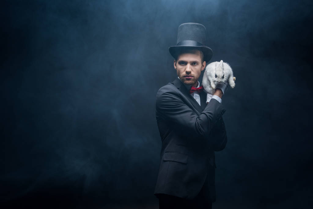 professional magician in suit and hat holding white bunny, dark room with smoke - Photo, Image