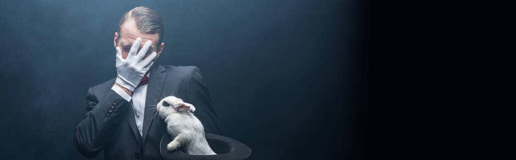 tiro panorâmico de emocional assustado mágico fechar rosto e segurando coelho branco em chapéu, sala escura com fumaça
 - Foto, Imagem