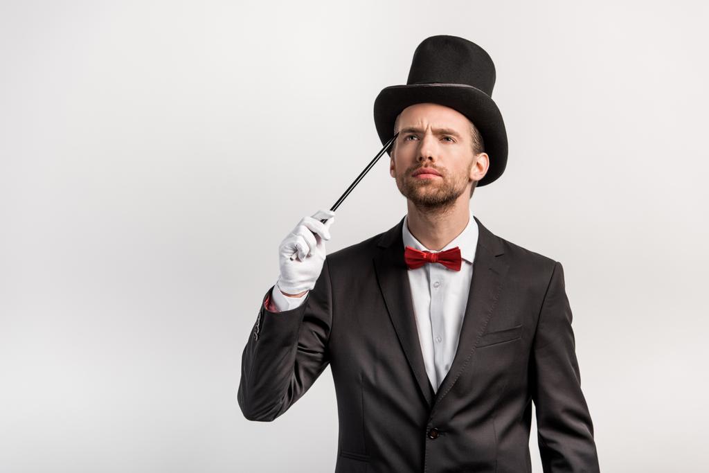 задумчивый фокусник в костюме и шляпе с палочкой, изолированный на сером
 - Фото, изображение
