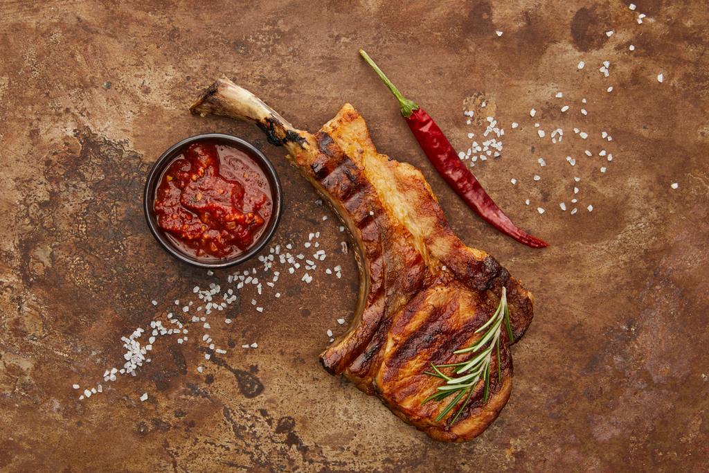 Horní pohled na ribeye steak s chilli paprikou, rajčatovou omáčkou a solí na kamenném pozadí - Fotografie, Obrázek