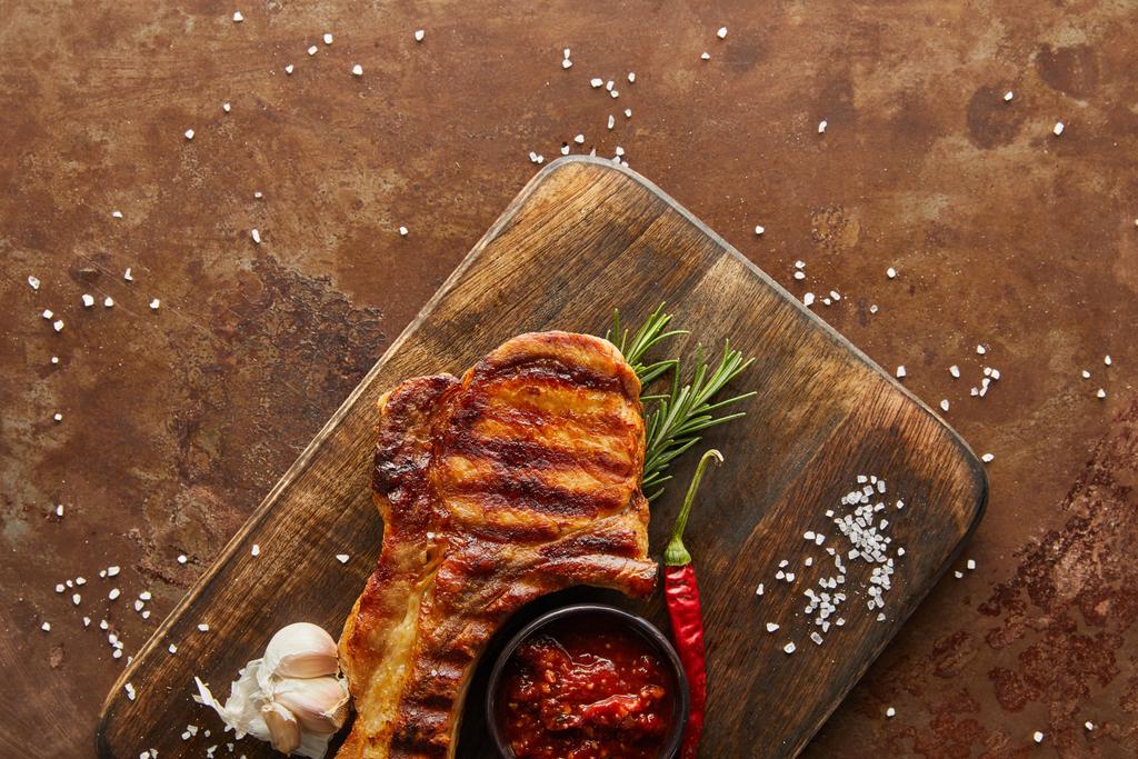 Horní pohled na chutný steak s chilli omáčkou, česnekem a rozmarýnem na řezací desce na kamenném pozadí - Fotografie, Obrázek
