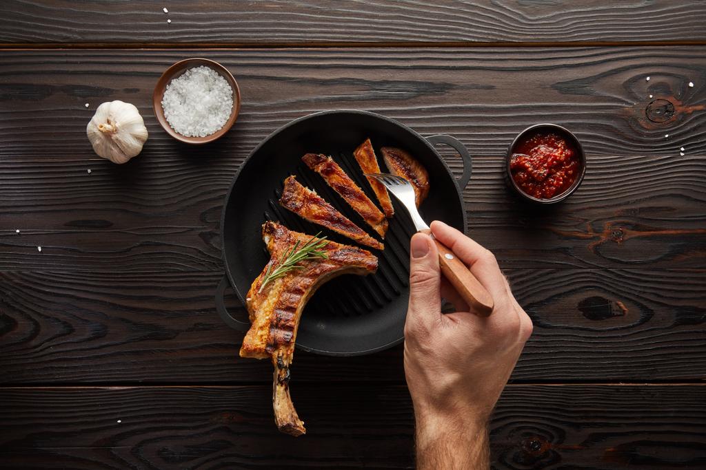 Top view of man holding fork with steak slice by tomato sauce, salt and garlic on wooden background - Photo, Image