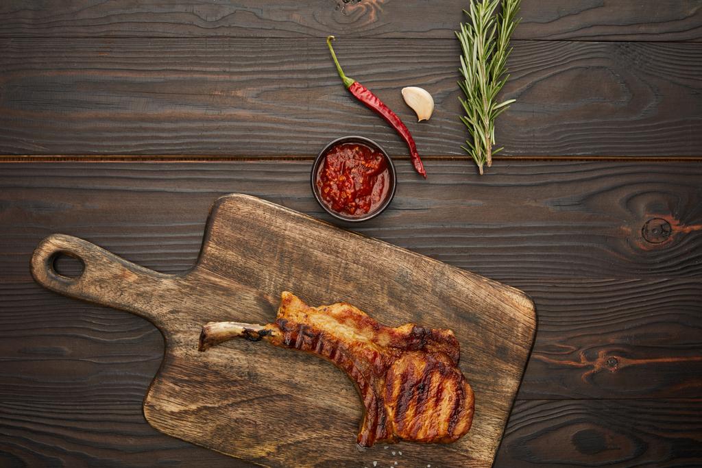 Top view of delicious ribeye steak on cutting board with chili sauce, rosemary and garlic on wooden background - Photo, Image