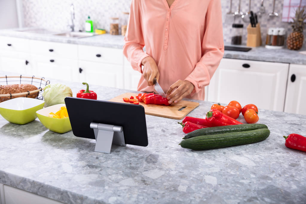 Donna che utilizza la tavoletta digitale mentre taglia il peperone rosso sul tagliere in cucina
 - Foto, immagini