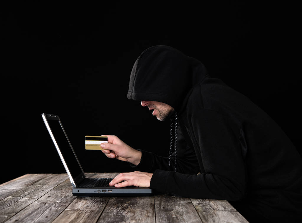 hacker v černé Hoody s notebookem a bankovní kartou před černým pozadím - Fotografie, Obrázek