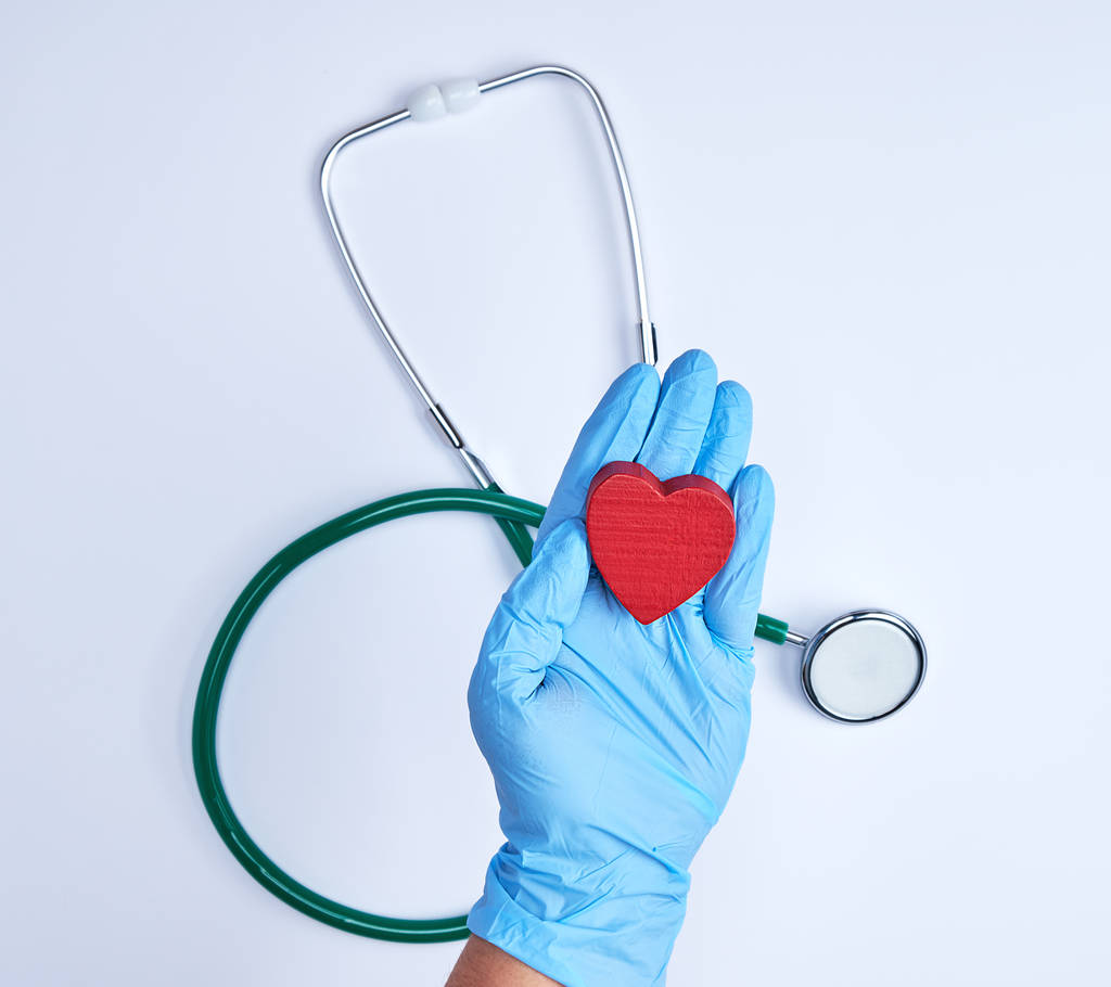 ανθρώπινο χέρι με μπλε αποστειρωμένα γάντια που κρατώντας μια κόκκινη καρδιά, λευκό φόντο με ένα στηθοσκόπιο, κάτοψη - Φωτογραφία, εικόνα