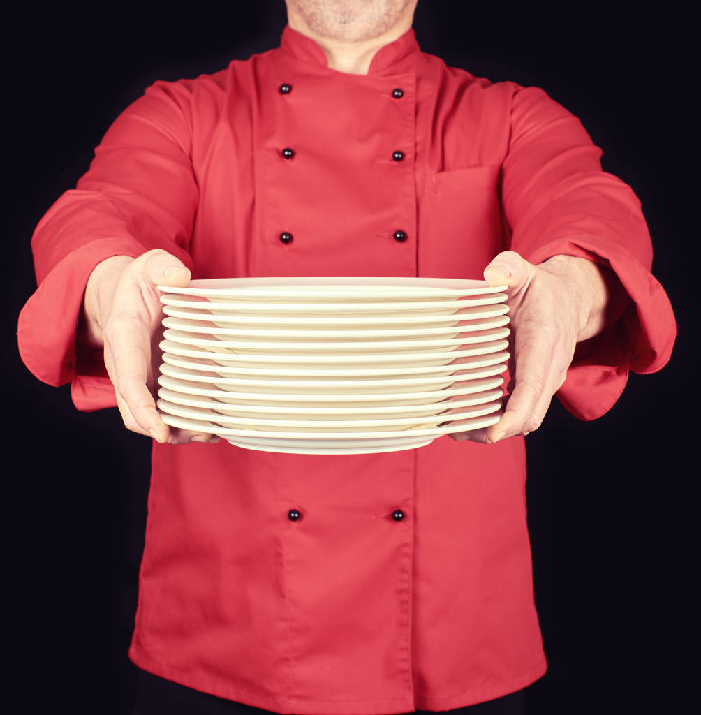 Cook a piros egységes tart a kezében egy rakás kerek fehér üres lemezek, fekete háttér - Fotó, kép