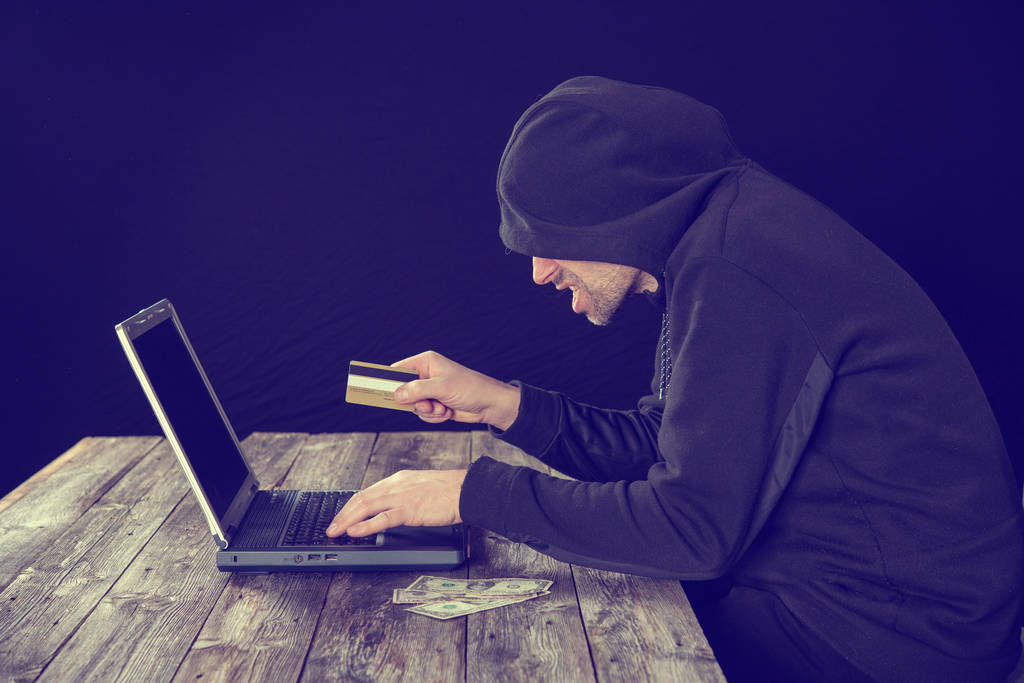 haker w czarnej bluzie z laptopem karty bankowej i banknotów dolarowych przed czarnym tle - Zdjęcie, obraz