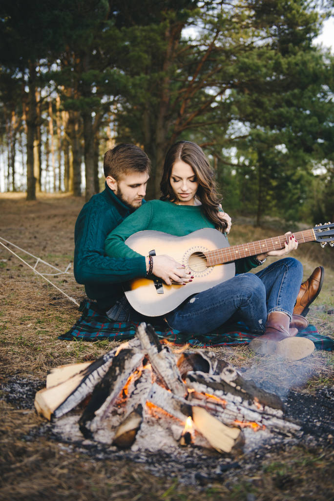 Pareja joven enamorada acampar turistas sentados junto a una fogata contra una tienda en el bosque tocando la guitarra
 - Foto, imagen