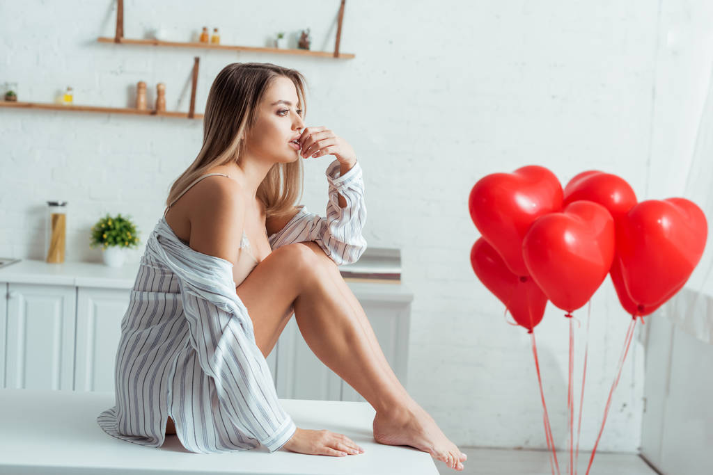 Seksi kadın masada oturuyor ve kırmızı kalp şeklinde balonların yanında dudaklarına dokunuyor.  - Fotoğraf, Görsel