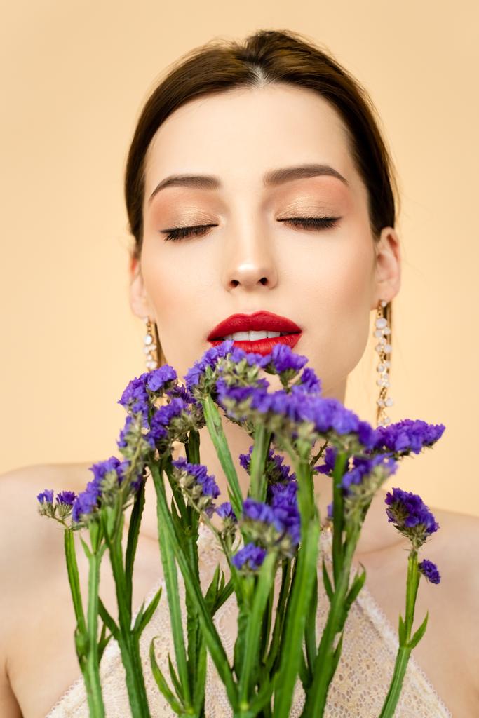 όμορφη γυναίκα με κλειστά μάτια κοντά σε μωβ λουλούδια limonium απομονωμένη σε μπεζ  - Φωτογραφία, εικόνα