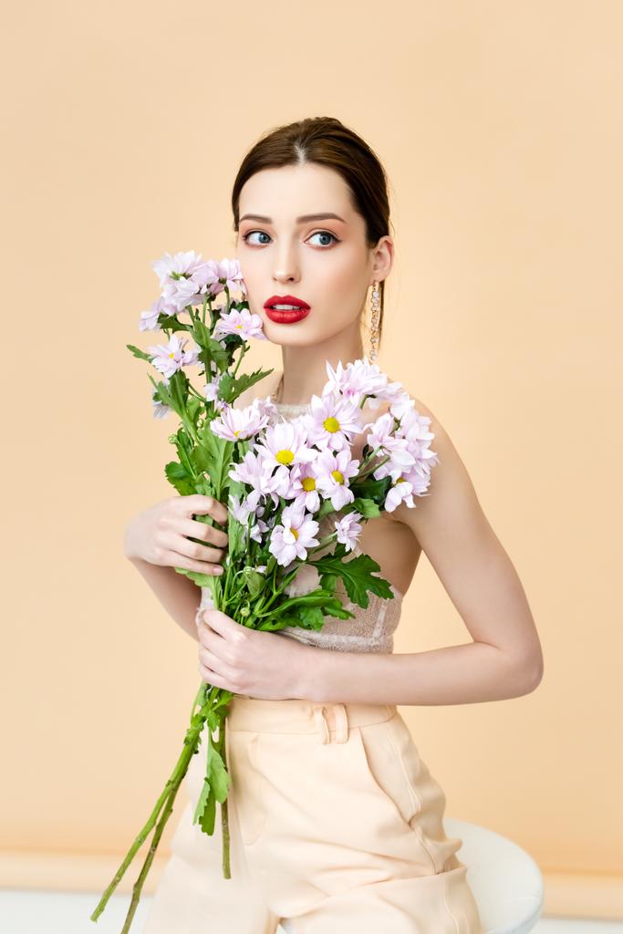 όμορφη γυναίκα κοιτάζει μακριά και κρατώντας ανθισμένα λουλούδια χρυσάνθεμα σε μπεζ  - Φωτογραφία, εικόνα