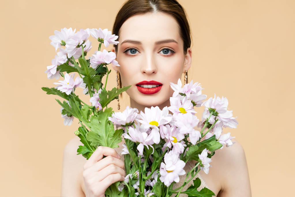 schönes Mädchen blickt in die Kamera und hält blühende Chrysanthemenblüten isoliert auf beige  - Foto, Bild