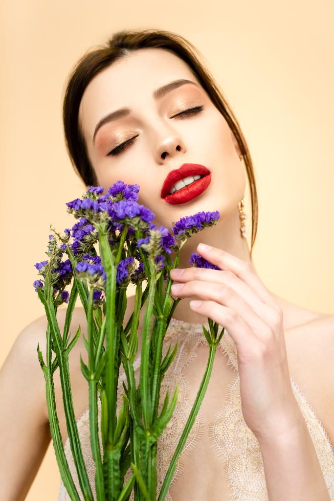 όμορφη γυναίκα με κλειστά μάτια κρατώντας μωβ λουλούδια limonium απομονωμένο σε μπεζ  - Φωτογραφία, εικόνα