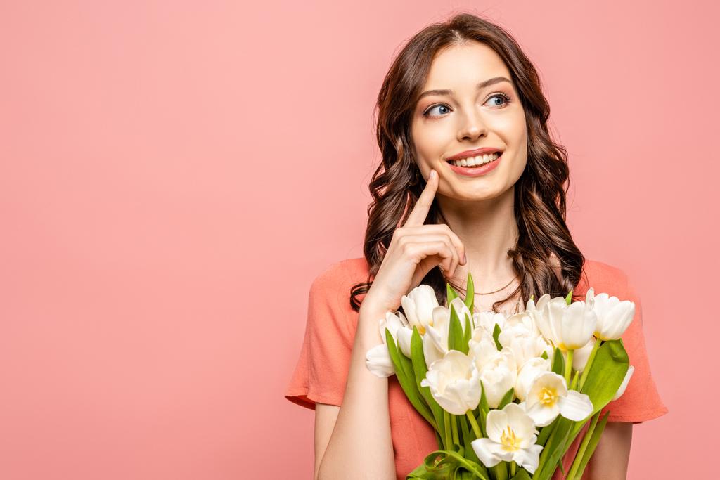 chica feliz mirando hacia otro lado y tocando la mejilla mientras sostiene ramo de tulipanes blancos aislados en rosa
 - Foto, imagen