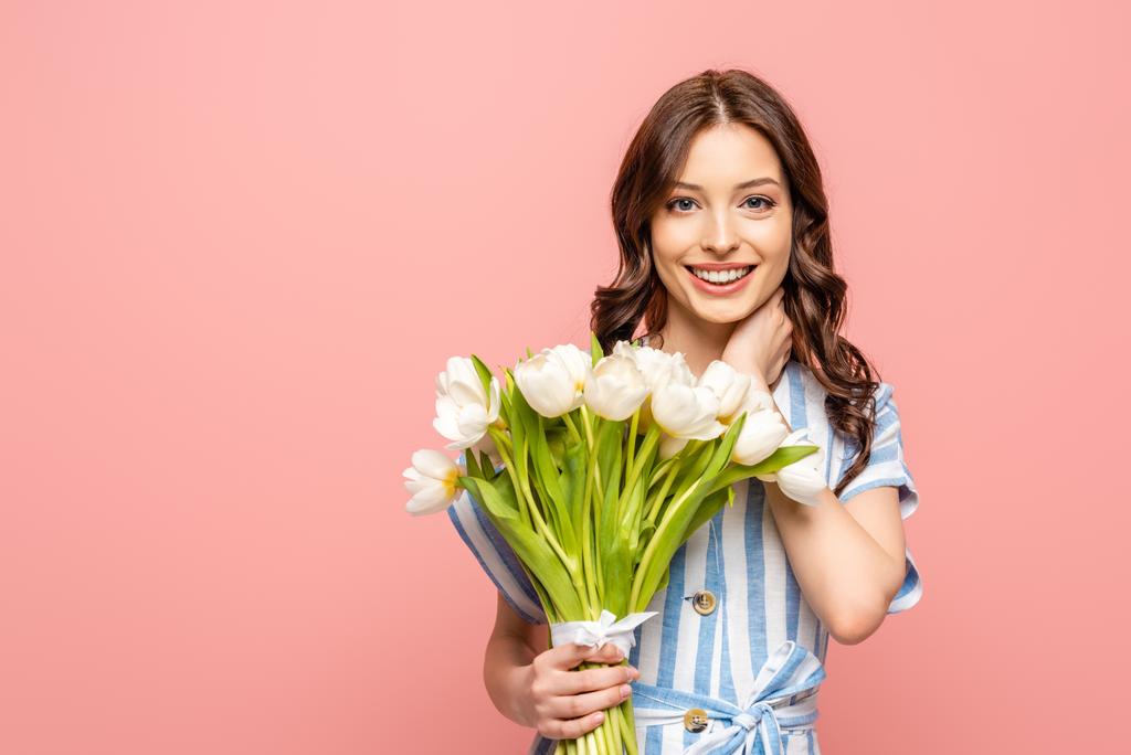 chica feliz tocando el cuello mientras sostiene ramo de tulipanes blancos y sonriendo a la cámara aislada en rosa
 - Foto, imagen