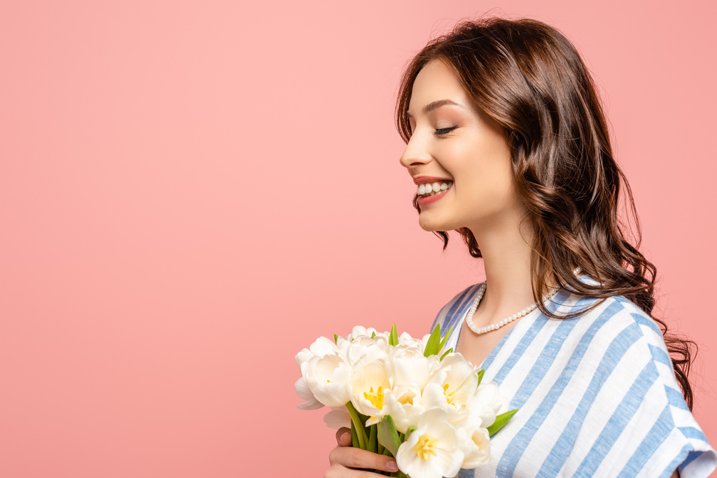 chica feliz sonriendo con los ojos cerrados mientras sostiene ramo de tulipanes blancos aislados en rosa
 - Foto, Imagen