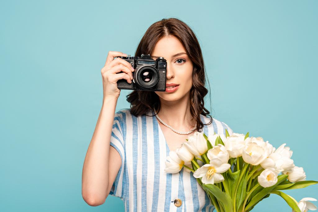 attraktive junge Frau fotografiert auf Digitalkamera, während sie einen Strauß weißer Tulpen auf blauem Grund hält - Foto, Bild