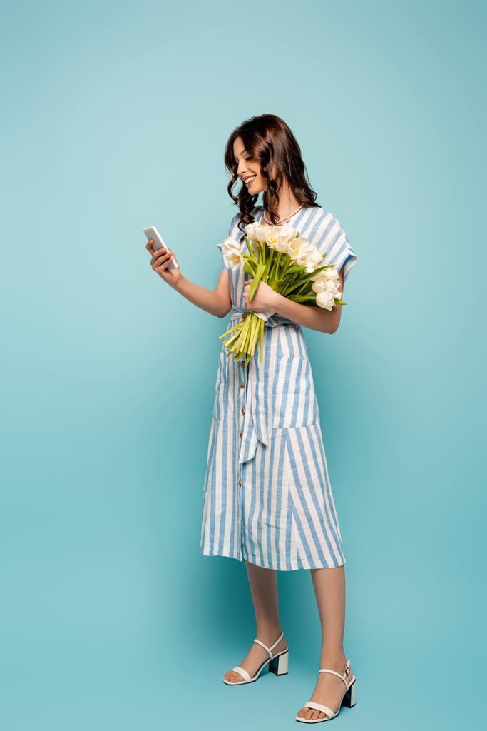 plná délka pohled na atraktivní mladá žena chatování na smartphone zatímco drží bílé tulipány na modrém pozadí - Fotografie, Obrázek
