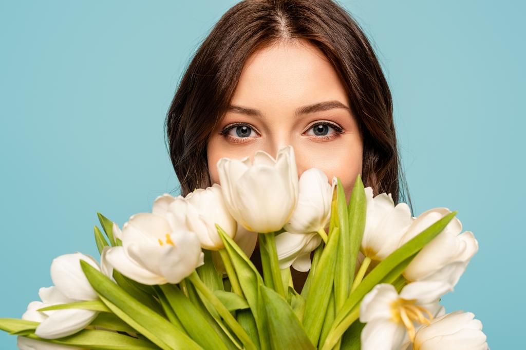 schöne junge Frau genießt Geschmack der weißen Tulpen, während sie isoliert auf blau in die Kamera schaut - Foto, Bild