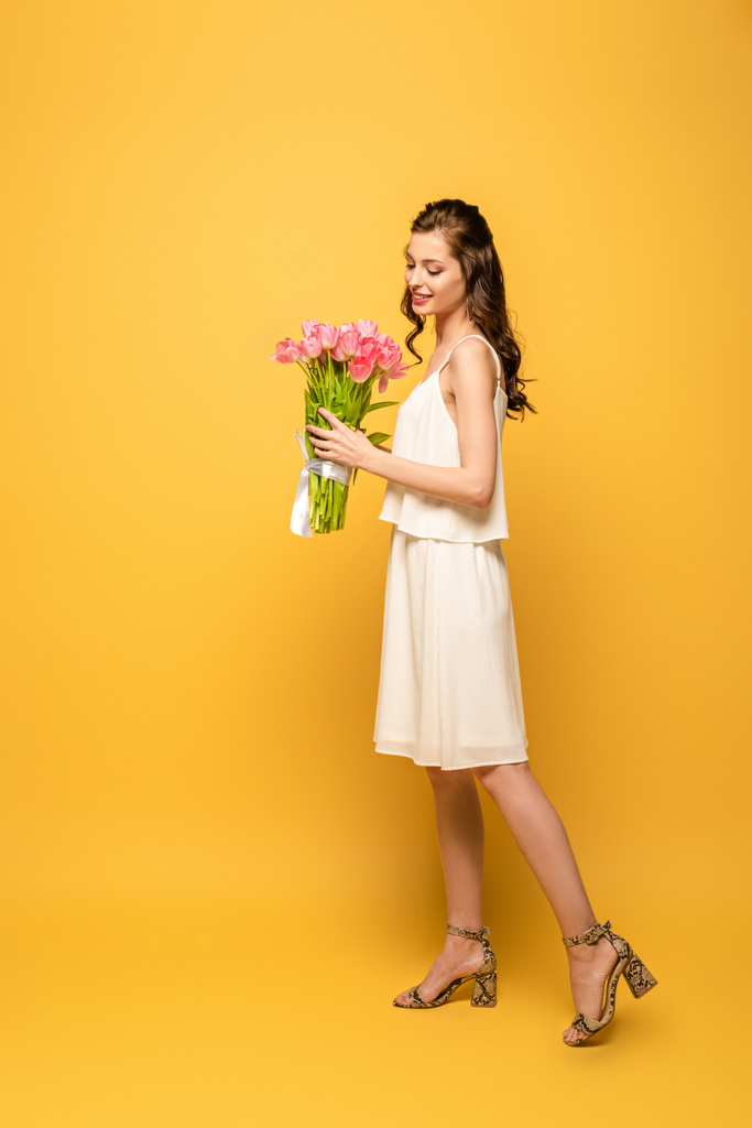 volledige weergave van glimlachende jonge vrouw met boeket van roze tulpen op gele achtergrond - Foto, afbeelding