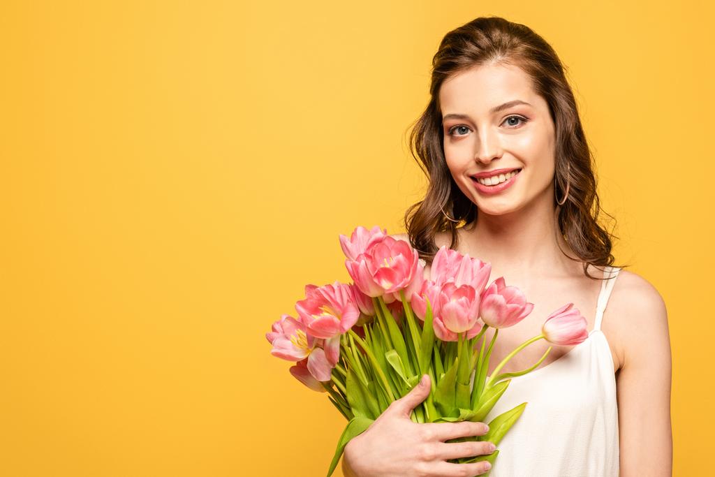 jovem mulher alegre sorrindo para a câmera enquanto segurando buquê de tulipas rosa isolado no amarelo
 - Foto, Imagem