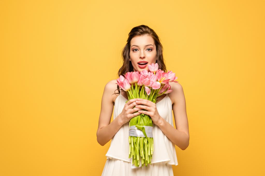 изумленная молодая женщина, держащая букет розовых тюльпанов, глядя на камеру, изолированную на желтый
 - Фото, изображение
