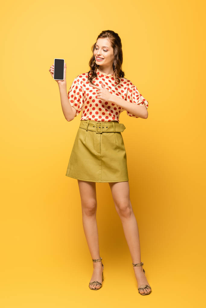 vista completa de la atractiva mujer joven sonriendo mientras señala con el dedo en el teléfono inteligente con pantalla en blanco sobre fondo amarillo
 - Foto, imagen