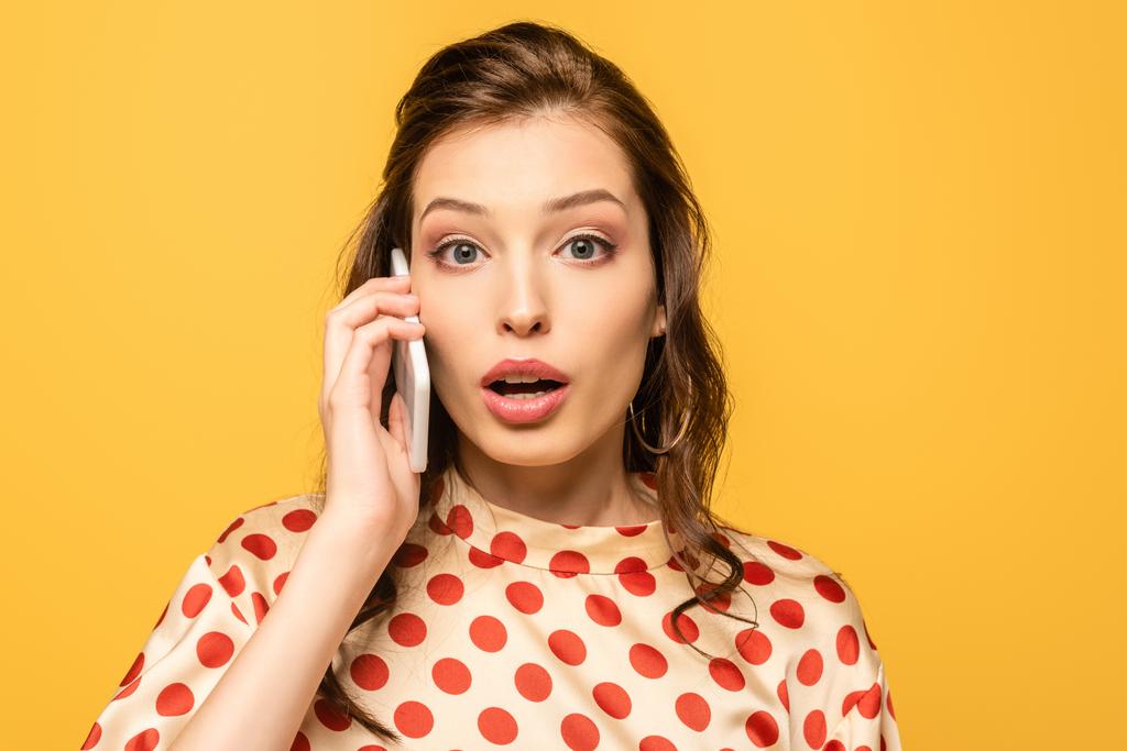 shocked young woman looking at camera while talking on smartphone isolated on yellow - Photo, Image