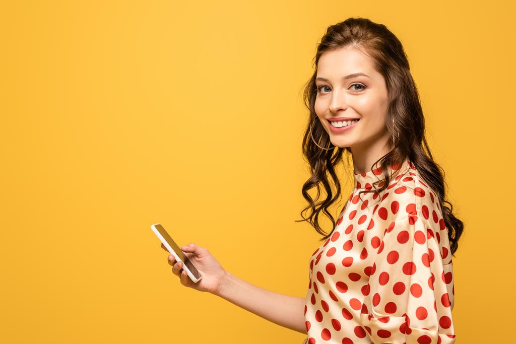 alegre joven mujer sosteniendo teléfono inteligente mientras sonríe a la cámara aislada en amarillo
 - Foto, imagen