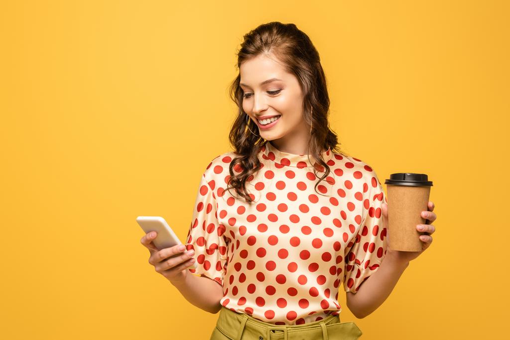 feliz mujer joven sosteniendo café para ir mientras se utiliza el teléfono inteligente aislado en amarillo
 - Foto, Imagen