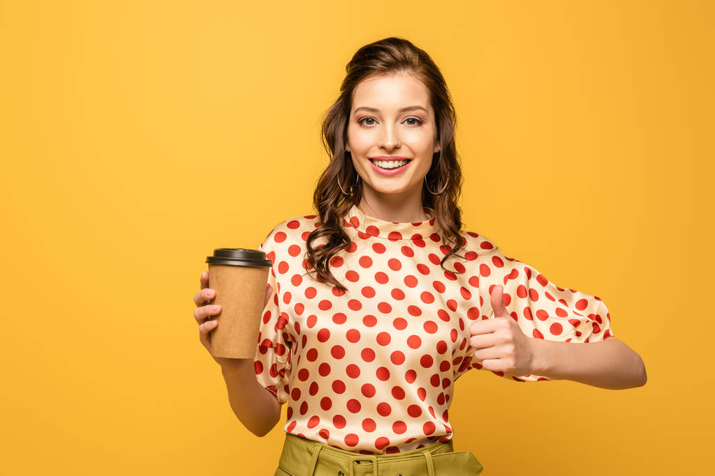 felice giovane donna che tiene il caffè per andare e mostrando pollice in su mentre guarda la fotocamera isolata sul giallo
 - Foto, immagini