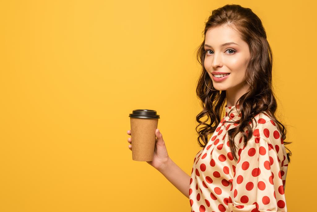 улыбающаяся молодая женщина держит кофе, чтобы пойти, глядя на камеру изолированы на желтый
 - Фото, изображение