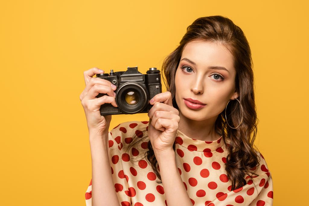 pewna siebie młoda kobieta trzyma aparat cyfrowy patrząc na aparat odizolowany na żółto - Zdjęcie, obraz