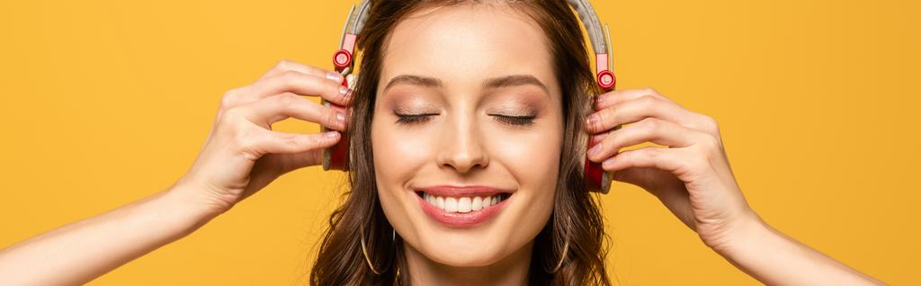 Panoramaaufnahme einer glücklichen jungen Frau, die Musik in drahtlosen Kopfhörern hört, mit geschlossenen Augen, isoliert auf gelb - Foto, Bild