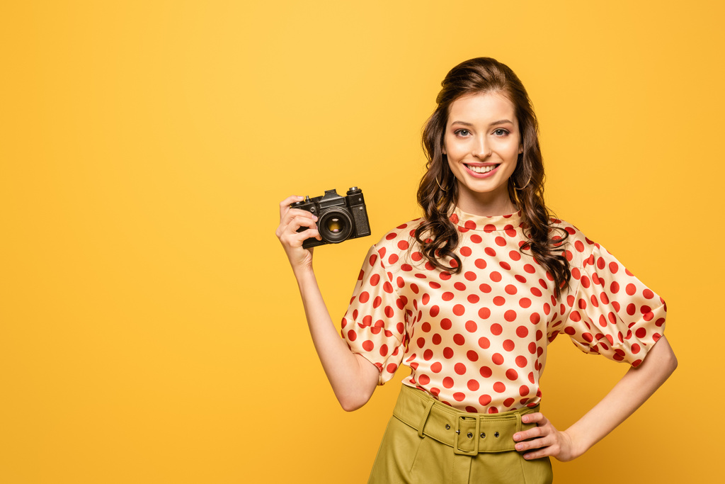 веселая молодая женщина держит цифровую камеру, стоя с рукой на бедре изолированы на желтый
 - Фото, изображение