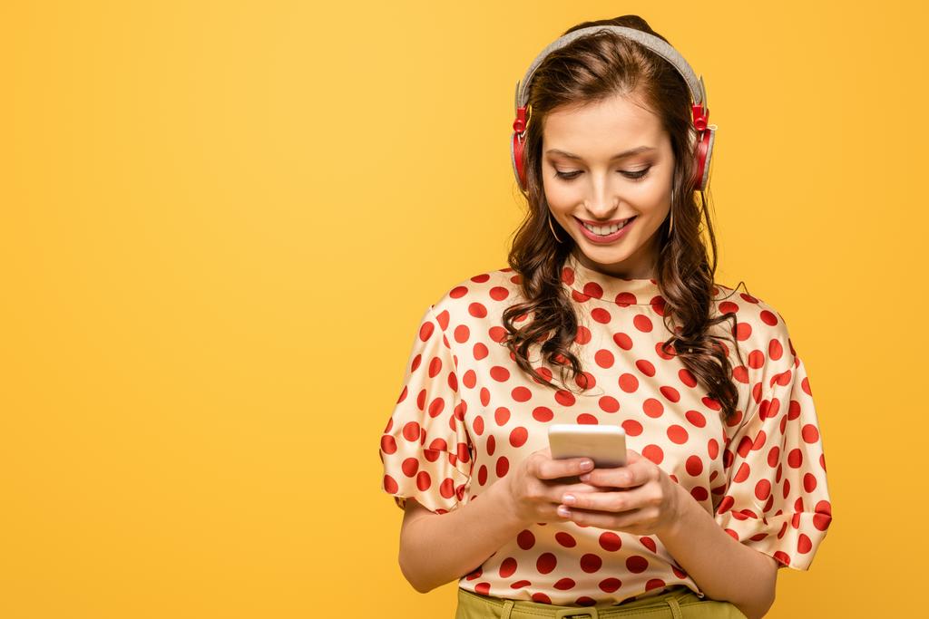 黄色で隔絶されたスマートフォンでおしゃべりするワイヤレスヘッドフォンの陽気な若い女性 - 写真・画像