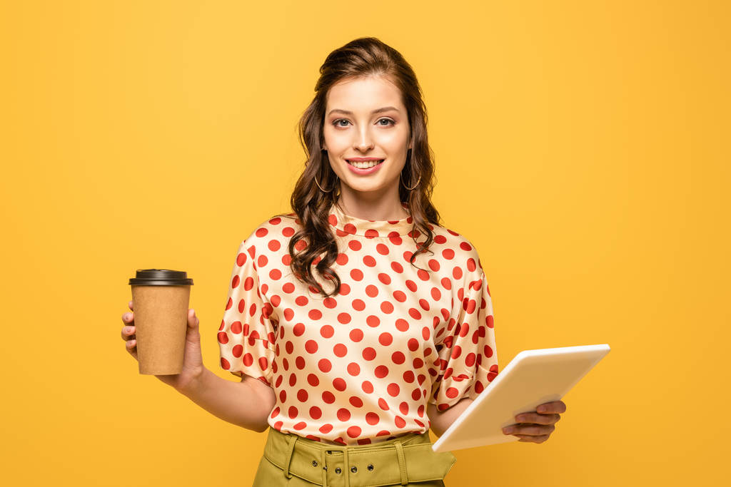 Ευτυχισμένη νεαρή γυναίκα χαμογελά στην κάμερα, ενώ κρατώντας τον καφέ για να πάει απομονωμένη στο κίτρινο - Φωτογραφία, εικόνα