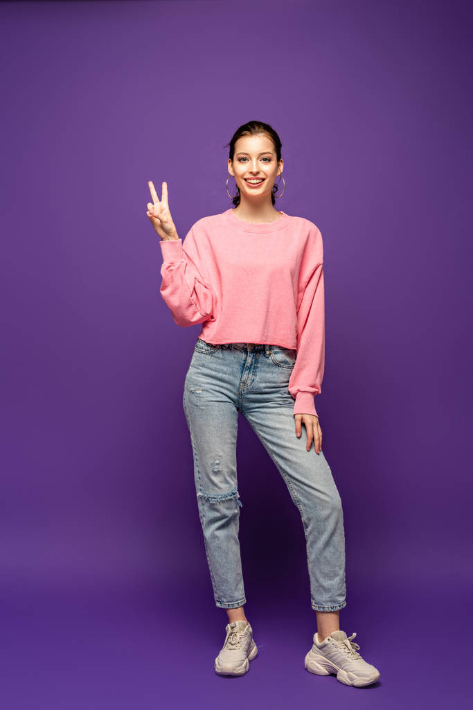 full length view of attractive girl smiling at camera while showing victory gesture on purple background - Photo, Image