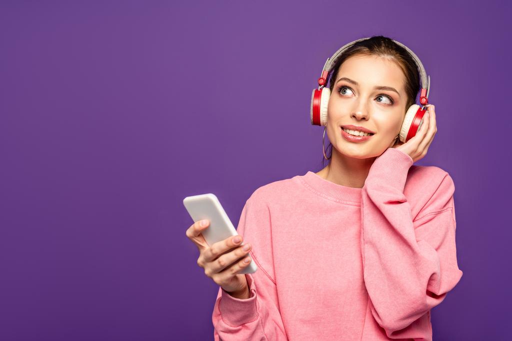 hymyilevä, unenomainen tyttö kuuntelee musiikkia langattomissa kuulokkeissa pitäen älypuhelinta ja katsoen pois eristettynä violettiin
 - Valokuva, kuva