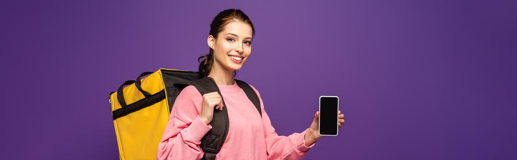 panoramiczne ujęcie ładnego kuriera niosącego termo box i trzymającego smartfona z pustym ekranem odizolowanym na fioletowym - Zdjęcie, obraz