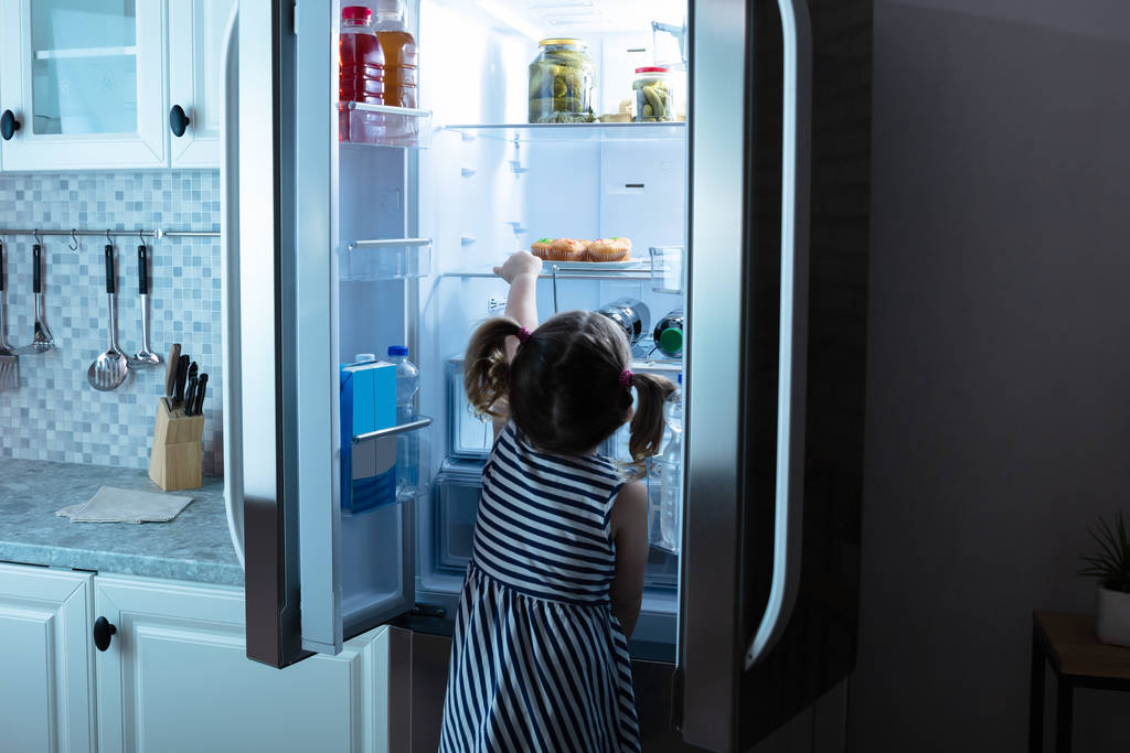 Visão traseira da menina tentar tirar muffins da geladeira na cozinha
 - Foto, Imagem