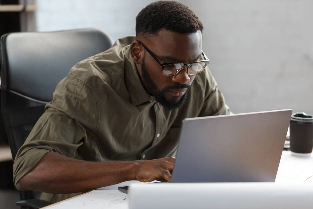 Afroamerikanischer Architekt, der im Büro mit Laptop arbeitet.Geschäftsporträt eines schwarzen gutaussehenden bärtigen Mannes mit Brille, der am Arbeitsplatz sitzt.Selbstbewusster Geschäftsmann wurde erfolgreich.Geschäftskonzept - Foto, Bild