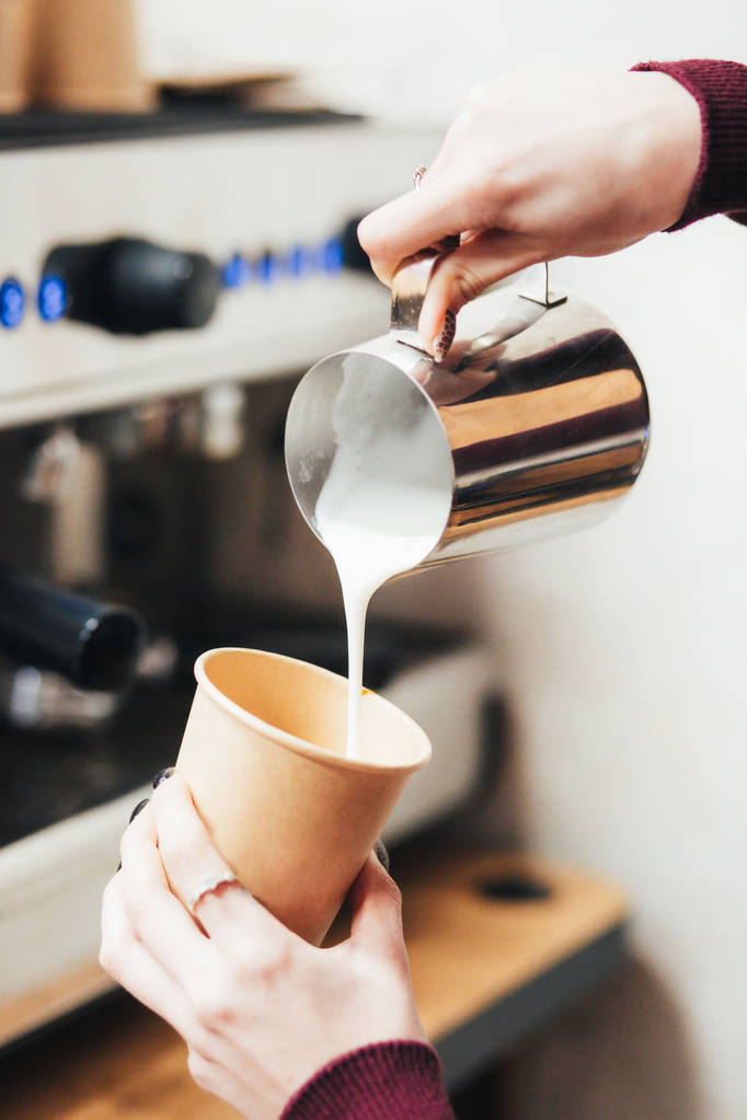 Ο Μπαρίστα ετοιμάζει καφέ. Ρίχνει γάλα από το βάζο σε ένα φλιτζάνι καφέ, καφέ που ετοιμάζεται από έναν barista. Εστίαση στα χέρια κρατώντας φλιτζάνι καφέ - Φωτογραφία, εικόνα