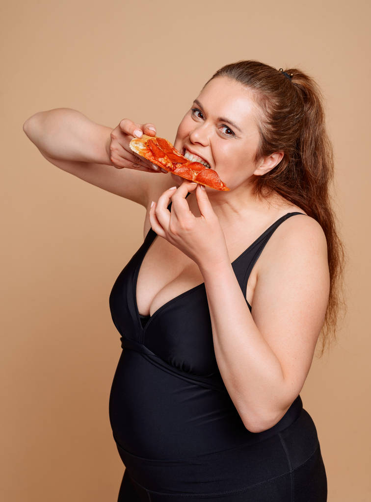 Nadváha žena jíst pizzu plátek dychtivě na béžové pozadí - Fotografie, Obrázek