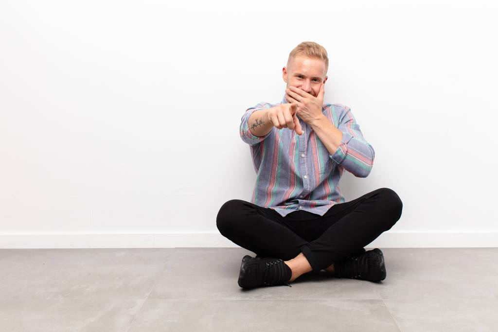young blonde man laughing at you, pointing to camera and making fun of or mocking you sitting on cement floor - Photo, Image