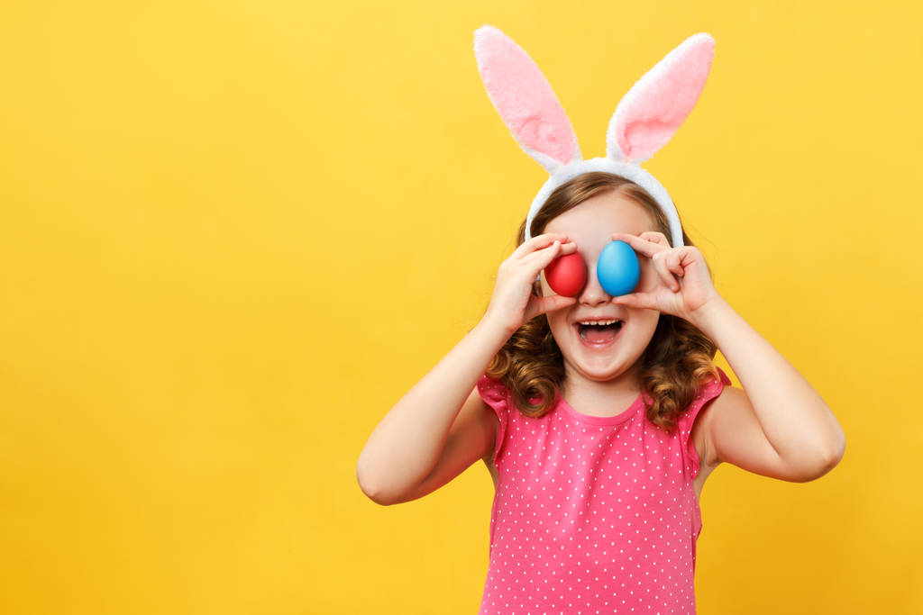 Wesołe, szczęśliwe dziecko w uszach króliczka trzyma w oczach dwa pisanki. Portret dziewczynki na żółtym tle w studiu - Zdjęcie, obraz