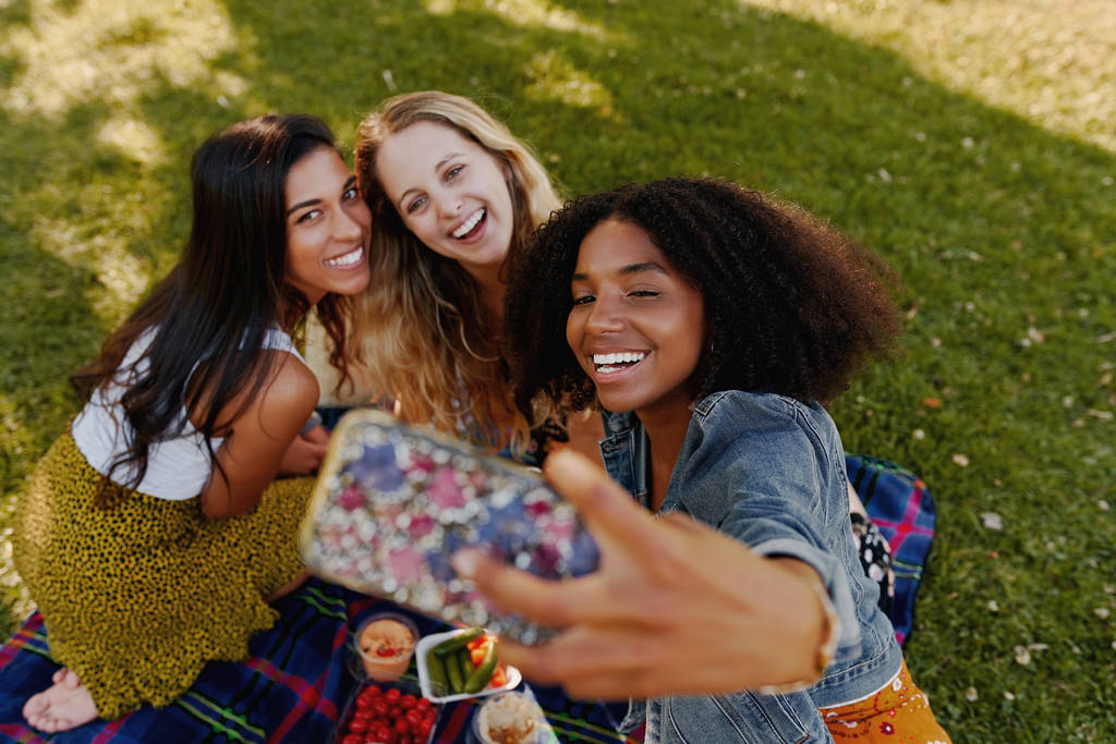 Widok wesołych wielorasowych przyjaciół robiących selfie przez telefon komórkowy na pikniku w parku - Zdjęcie, obraz