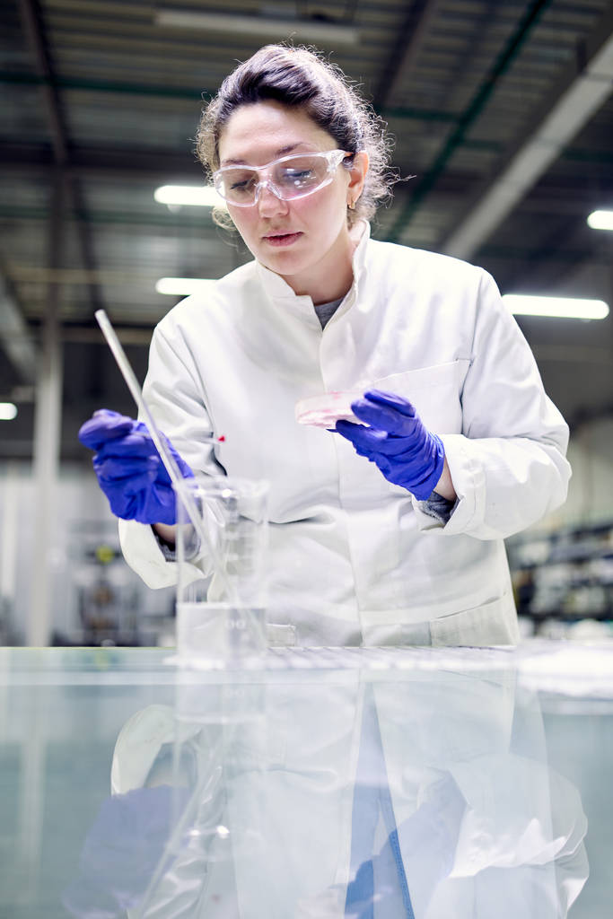 Jovem mulher de laboratório em óculos e placa de Petri em suas mãos realiza experimentos
 - Foto, Imagem